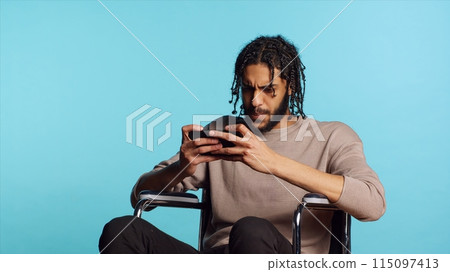 Gamer with disability upset after receiving game over screen on smartphone display. BIPOC man in wheelchair feeling sad after losing videogame, playing on mobile phone, camera A 115097413