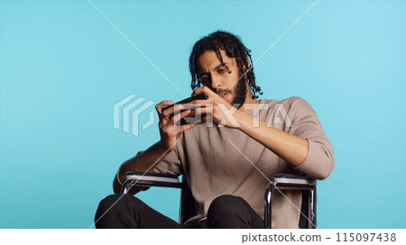 Man with disability in wheelchair playing videogames on cellphone, enjoying leisure time. Middle Eastern gamer enjoying game on mobile phone, having fun, studio background, camera A Man with disability in wheelchair playing videogames on cellphone, enjoying leisure time. Middle Eastern gamer enjoying game on mobile phone, having fun, studio background, camera A 115097438