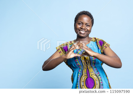 Young person creating heart shape gesture in the studio, expressing her sincere emotions with a romantic symbol. African american woman feeling optimistic, true love and feelings. Young person creating heart shape gesture in the studio, expressing her sincere emotions with a romantic symbol. African american woman feeling optimistic, true love and feelings. 115097590