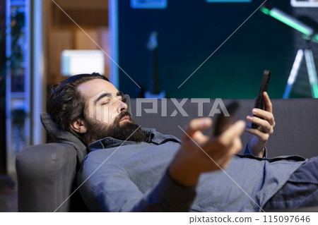Homeowner bored at home laying down, using TV to watch movies on streaming services and chatting with friends. Apathetic man lounged on couch, mindlessly consuming entertainment and using mobile phone 115097646
