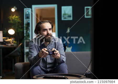 Joyous man playing videogame in home theatre, holding controller. Smiling gamer participating in PvP online multiplayer game using console system and high quality gamepad 115097664