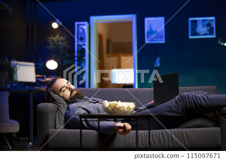 Man sleeping on couch from tiredness after working on laptop all day before managing to watch TV and enjoy bowl of popcorn. Freelancer falls asleep while solving tasks on notebook and looking at film Man sleeping on couch from tiredness after working on laptop all day before managing to watch TV and enjoy bowl of popcorn. Freelancer falls asleep while solving tasks on notebook and looking at film 115097671