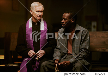 African American man holding rosary sitting on pew having conversation with senior Caucasian Catholic priest, medium shot African American man holding rosary sitting on pew having conversation with senior Caucasian Catholic priest, medium shot 115105221