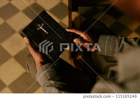 High angle view of hands of unrecognizable Black man sitting on pew in Catholic church holding prayer book High angle view of hands of unrecognizable Black man sitting on pew in Catholic church holding prayer book 115105249