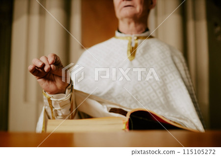 Selective focus shot of senior Catholic pastor wearing vestment praying while standing at lectern with book on it 115105272