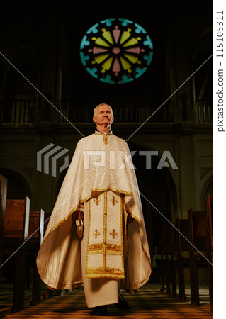 Vertical low angle long shot of senior Caucasian Catholic priest wearing liturgical vestment walking along church nave Vertical low angle long shot of senior Caucasian Catholic priest wearing liturgical vestment walking along church nave 115105311