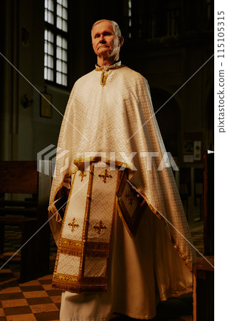Vertical low angle full length shot of senior Catholic priest wearing liturgical clothes walking along church nave and looking at camera 115105315