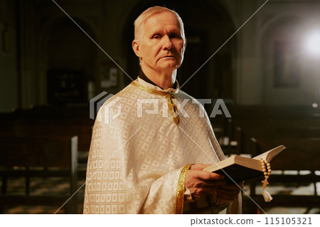 Medium portrait of elderly Catholic priest wearing beige chasuble standing indoors with Bible in hands looking at camera 115105321
