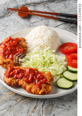 Ayam geprek marinated chicken that's pounded, deep-fried and finished with a spicy sambal served with white rice, cucumber, tomato and cabbage closeup on the plat. Vertical 115109938