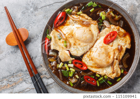 Fried eggs in soy sauce with onions, green onions, chili peppers and garlic close-up in a plate on the table. Horizontal top view 115109941