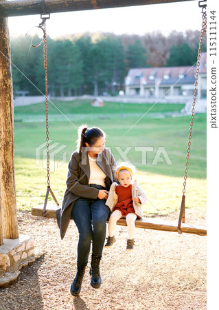 Mom hugs a little girl sitting with her on a wooden chain swing in the park 115111144