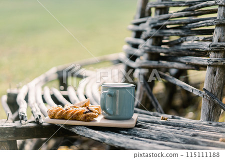 Mug of coffee next to a bun stands on a wooden plank on a park bench Mug of coffee next to a bun stands on a wooden plank on a park bench 115111188