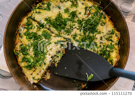 Omelet with greens and cheese in a pan with a spatula is on the table. Top view Omelet with greens and cheese in a pan with a spatula is on the table. Top view 115111270