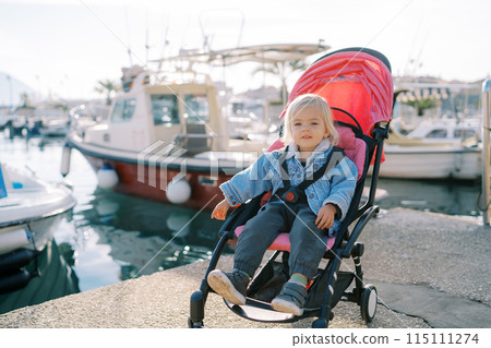 Little smiling girl sits in a stroller on the pier near the moored yachts and points to the sea Little smiling girl sits in a stroller on the pier near the moored yachts and points to the sea 115111274