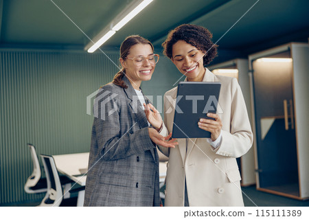 Stylish young female coworkers discussing project standing in modern office and use digital tablet Stylish young female coworkers discussing project standing in modern office and use digital tablet 115111389