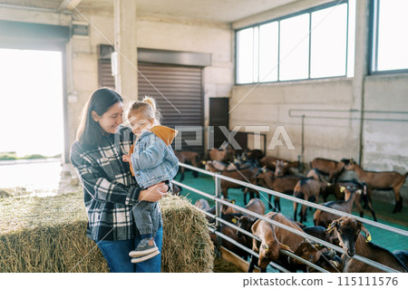Mom with a little girl in her arms stands by a haystack near a goat paddock Mom with a little girl in her arms stands by a haystack near a goat paddock 115111576