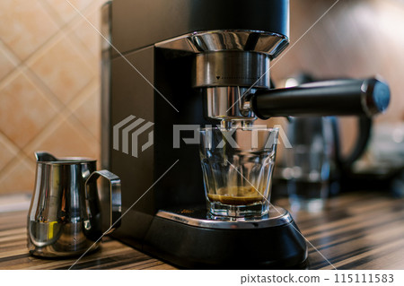 Black coffee drips into a glass on the tray of a carob coffee machine on a table Black coffee drips into a glass on the tray of a carob coffee machine on a table 115111583