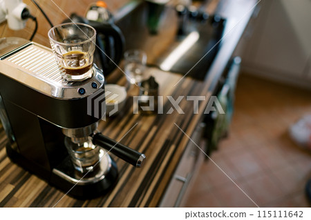 Glass of coffee stands on a carob coffee machine preparing coffee in a cup Glass of coffee stands on a carob coffee machine preparing coffee in a cup 115111642