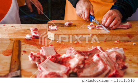 butcher cutting raw meat in to small pieces on the wooden board. ribs and ax on the foreground. table in blood. preparation for outdoor cooking of hungarian bogracs goulash in stew 115113516