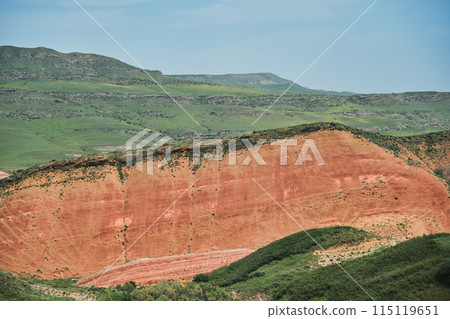 A breathtaking ethereal landscape with vibrant multi-colored red hills under a cloudy sky. A lush green valley stretches into the distance offering stunning views of nature's beauty 115119651