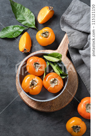 Fresh ripe persimmon fruits in bowl on a cutting board on a dark background with slices and leaves. Fresh ripe persimmon fruits in bowl on a cutting board on a dark background with slices and leaves. 115121150