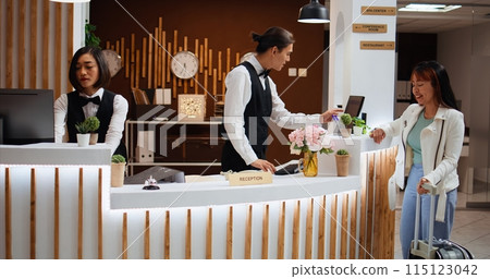 Hotel guest arriving at reception to do check in process, claiming room reservation after long flight. Woman traveler on vacation at five star resort talking to receptionist about facilities. Hotel guest arriving at reception to do check in process, claiming room reservation after long flight. Woman traveler on vacation at five star resort talking to receptionist about facilities. 115123042