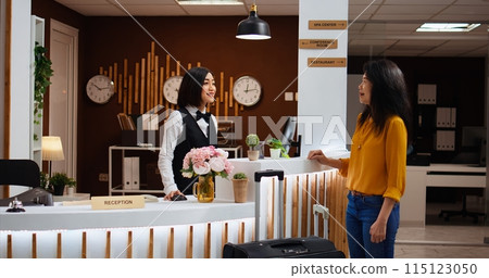 Receptionist welcoming asian woman at five star resort, asking about booking details to fill in registration forms. Front desk staff providing great services for client, presenting hotel amenities. 115123050