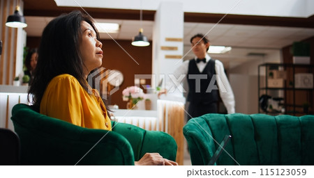 Hotel client solving issues on laptop in lounge area, waiting to register and see accommodation. Asian tourist passing time on pc, waiting to be called at front desk reception. Handheld shot. 115123059