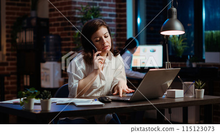 Woman answers phone call from husband while working in office at computer desk. Employee using smartphone to chat with friend during telephone call while at work in front of laptop at night, camera B 115123073