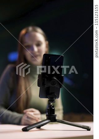 Focus on smartphone on tripod used by cheerful girl in blurry background to film in living room used as professional studio. Close up shot of phone used by teenager to record vlog 115123155