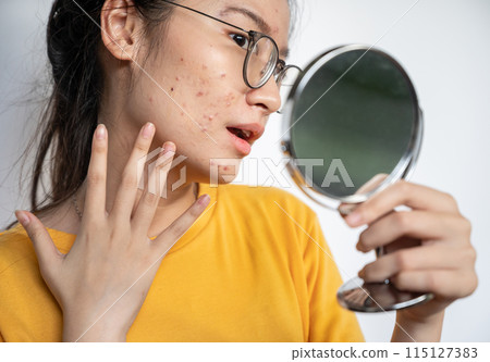 Young Asian woman feeling shock while looking acne and scar occur on face by mini mirror. Conceptual shot of Acne and Problem Skin on female face. 115127383