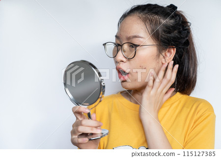 Young Asian woman feeling shock while looking acne and scar occur on face by mini mirror. Conceptual shot of Acne and Problem Skin on female face. 115127385