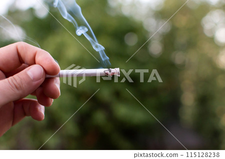 smoldering smoking cigarette in hand on the background of nature smoldering smoking cigarette in hand on the background of nature 115128238