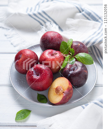 a few juicy red plums in plate with green leaves and napkin on a white wood background a few juicy red plums in plate with green leaves and napkin on a white wood background 115132056
