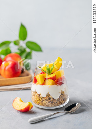 Yogurt parfait with peach and granola in a glass on a light background with fresh fruits. 115133219