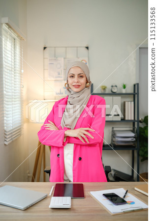 A woman in a pink jacket stands in front of a desk with a laptop, a cell phone A woman in a pink jacket stands in front of a desk with a laptop, a cell phone 115133568
