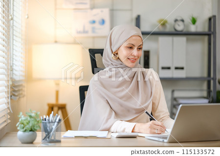A woman wearing a head scarf is sitting at a desk with a laptop and a notebook A woman wearing a head scarf is sitting at a desk with a laptop and a notebook 115133572