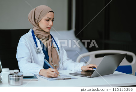 Female in uniform of doctor using digital technology laptop for Output Device and writing a patient report on the office desk. 115135396