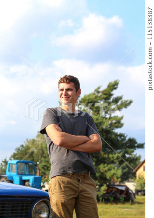 Outdoor portrait of a young Caucasian man in his 20s. Young handsome man with a happy smile, arms crossed, standing next to a car or tractor 115138387