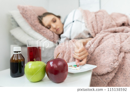On the bed lies a teenager and in front of her stands a table with apples and medicines that must be swallowed. On the bed lies a teenager and in front of her stands a table with apples and medicines that must be swallowed. 115138592