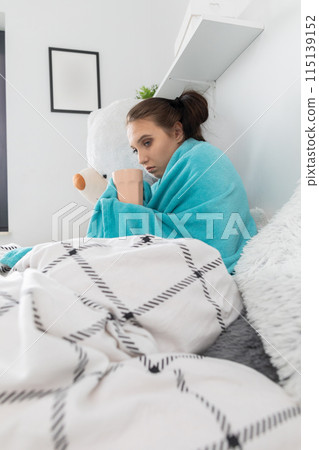 A young teenager drinks hot tea with herbs while lying on her bed under the covers. A young teenager drinks hot tea with herbs while lying on her bed under the covers. 115139152