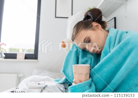 A young teenager drinks hot tea with herbs while lying on her bed under the covers. A young teenager drinks hot tea with herbs while lying on her bed under the covers. 115139153