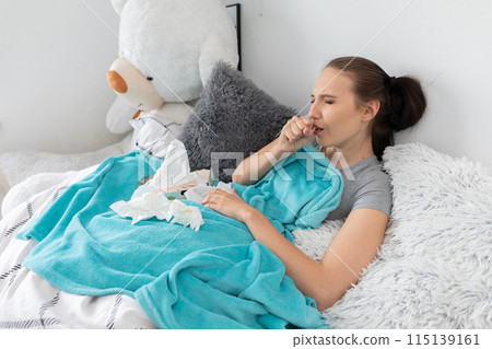 A teenager coughing strongly, cutting her mouth with her hand, lying in her bed under a warm blanket. A teenager coughing strongly, cutting her mouth with her hand, lying in her bed under a warm blanket. 115139161
