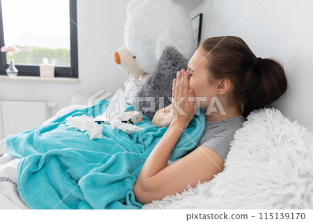 A teenager coughing strongly, cutting her mouth with her hand, lying in her bed under a warm blanket. A teenager coughing strongly, cutting her mouth with her hand, lying in her bed under a warm blanket. 115139170