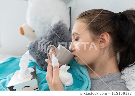 A young teenager drinks hot tea with herbs while lying on her bed under the covers. 115139182
