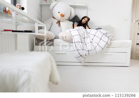 A sick teenager lies under the covers in bed next to her teddy bear. 115139266