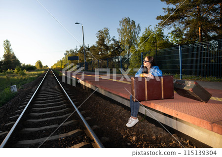 A teenager is sitting in a forbidden place on a railway platform and her legs are hanging over the railway tracks. Waiting for a late train. 115139304
