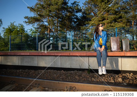 A teenager is sitting in a forbidden place on a railway platform and her legs are hanging over the railway tracks. Waiting for a late train. 115139308