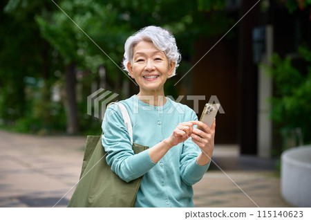 A cheerful senior woman holding a shopping bag and operating a smartphone 115140623