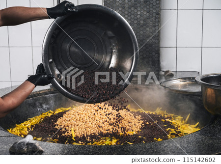 male chef's hands add raisins to cauldron with chickpeas and yellow carrots to cook traditional Uzbek pilaf in restaurant in Tashkent in Uzbekistan male chef's hands add raisins to cauldron with chickpeas and yellow carrots to cook traditional Uzbek pilaf in restaurant in Tashkent in Uzbekistan 115142602